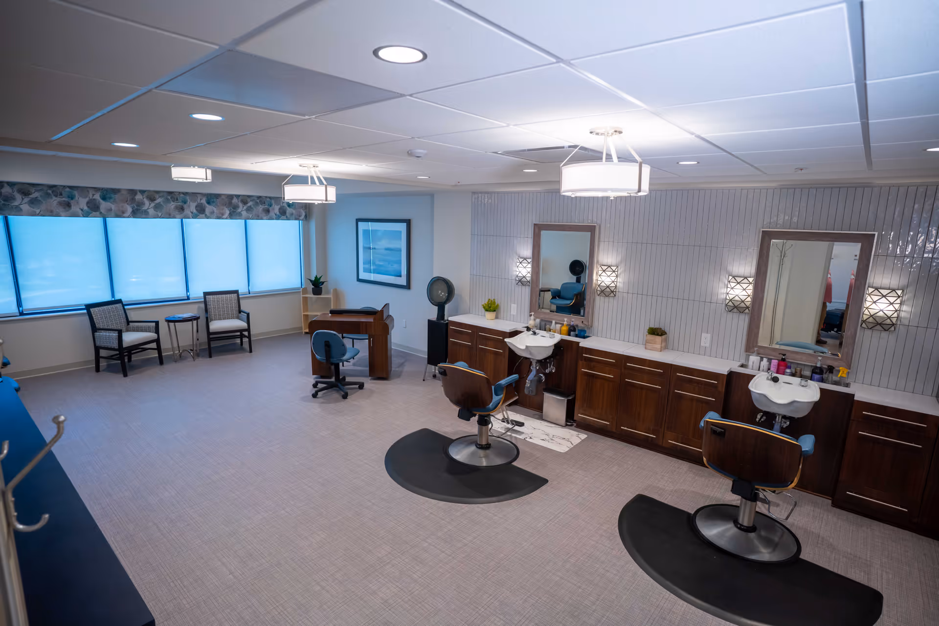 A spacious and well-lit salon area in a retirement community featuring two salon chairs with sinks, large mirrors, wooden cabinetry, and modern light fixtures. There are two chairs and a small table near large windows with blue shades, and a desk with an office chair in the corner.