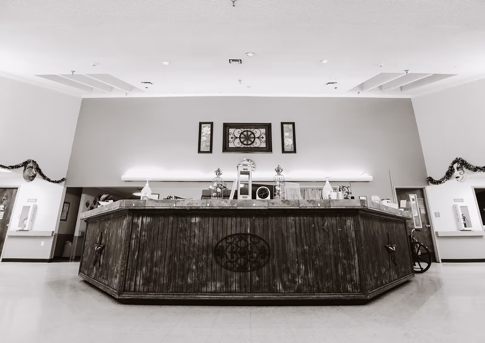 Black and white image of a large wooden reception desk in a spacious interior area with decorative wall art above it and garland decorations on the walls. The area appears clean and well-lit with a high ceiling and doors on either side of the desk.