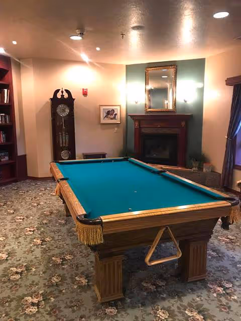 A cozy room with a pool table in the center, a grandfather clock against the wall, a fireplace with a mirror above it, and a bookshelf filled with books. The room has floral carpet and soft lighting.