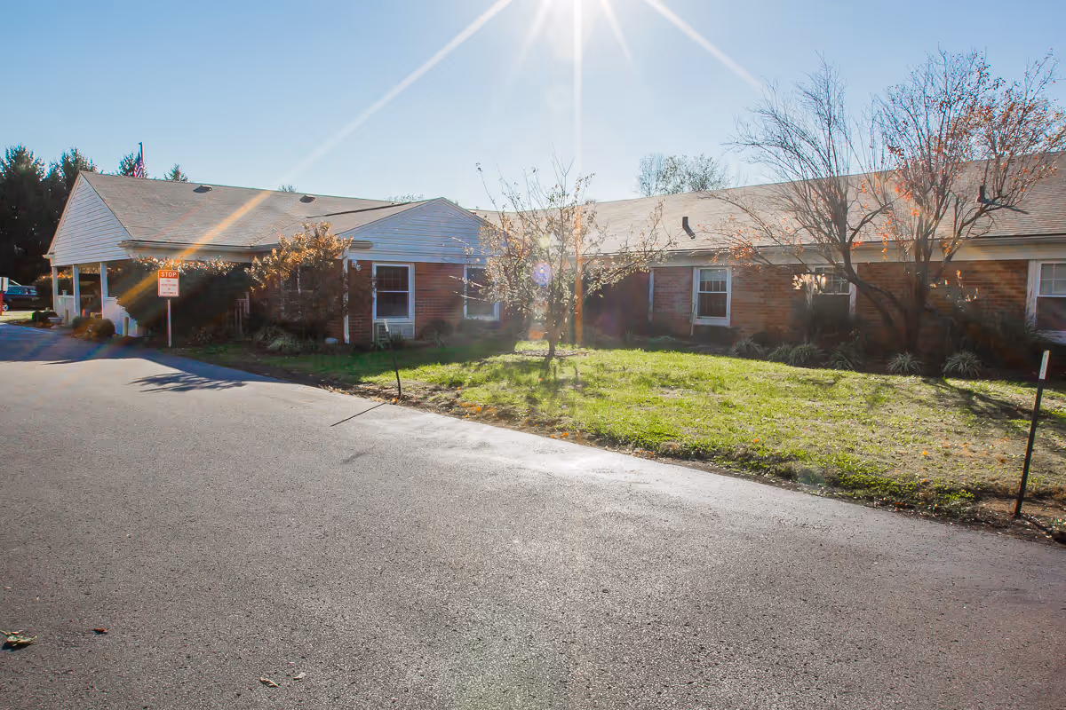 Front exterior of a single-story brick senior living facility with a driveway, lawn, and bright sun glare.