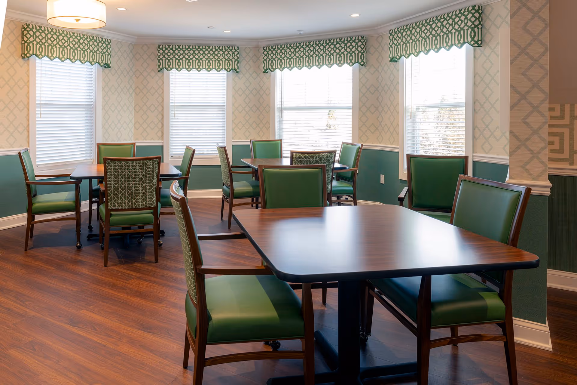 A dining room with multiple wooden tables and green cushioned chairs. The room has large windows with white blinds and green patterned valances. The walls are decorated with a geometric wallpaper pattern and a green wainscoting. The floor is a polished wood finish and a round ceiling light fixture is visible.