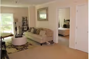 A cozy living room with a beige sofa adorned with patterned cushions, a round white ottoman, a floor lamp, and a decorative mirror on the wall. There is an open doorway leading to a bedroom with a bed visible. The room has beige carpeting and light-colored walls with a window letting in natural light.