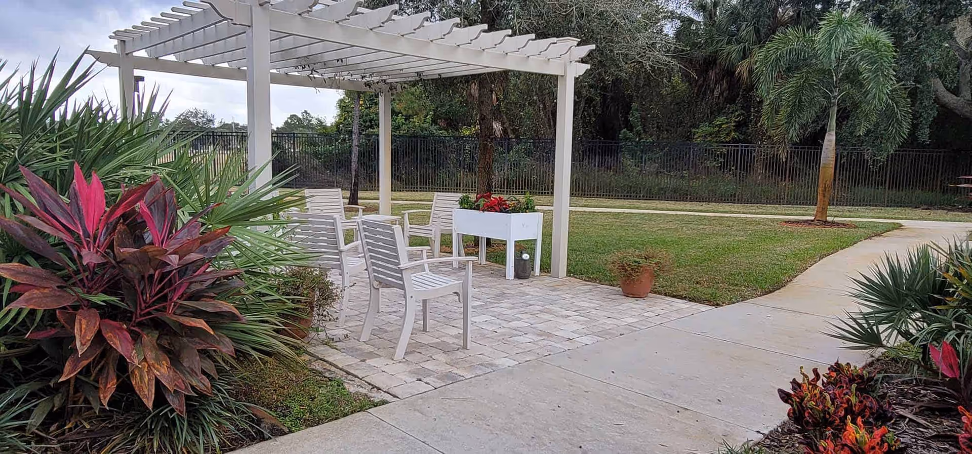 Outdoor patio with a white pergola, several white chairs, a planter and surrounding landscaping next to a paved walkway.