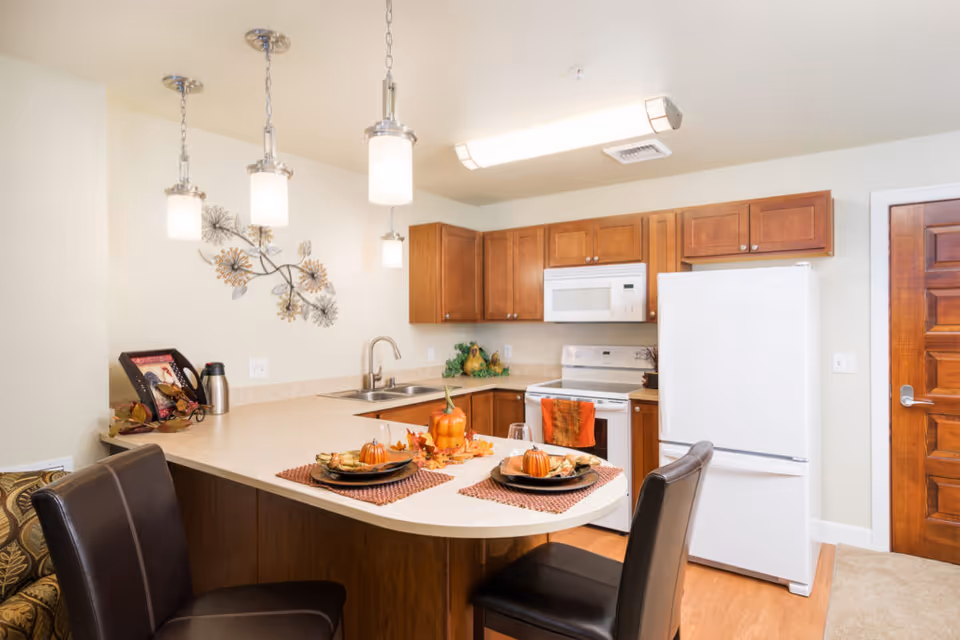 Bright kitchen with an island and two chairs, pendant lights, wooden cabinets, white appliances, and fall-themed place settings.