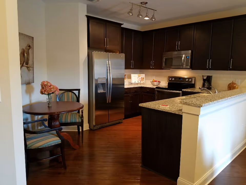 A modern kitchen with dark wood cabinets, stainless steel refrigerator, stove, microwave, and coffee maker. There is a granite countertop island with a sink and a small round dining table with two striped upholstered chairs and a vase of flowers. A painting of birds hangs on the wall near the dining area.