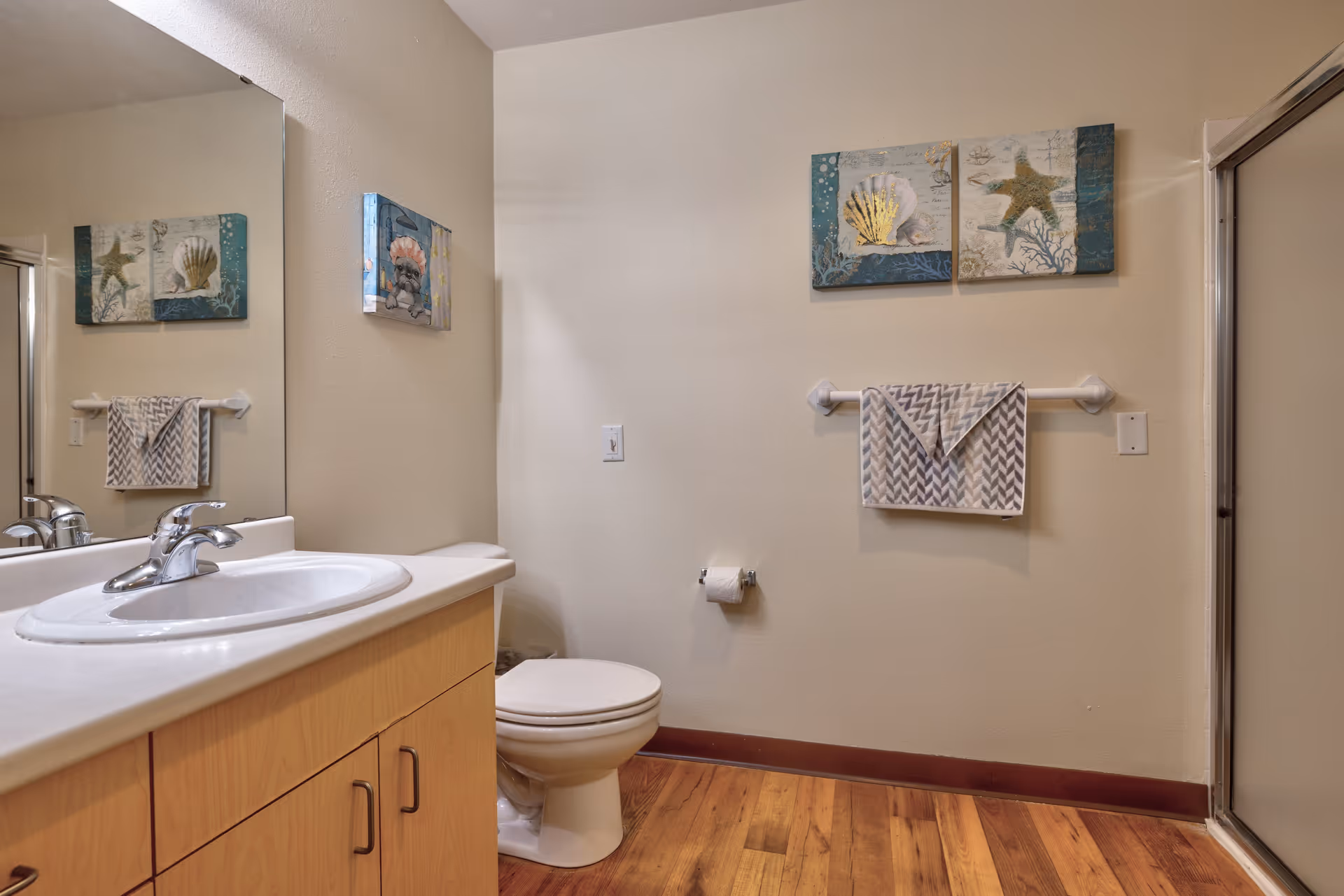 A bathroom with a wooden floor, a white sink with a silver faucet on a light wood cabinet, a toilet, a towel rack with a patterned towel, and two framed sea-themed artworks on the wall. There is a large mirror above the sink and a shower with a glass door on the right side.
