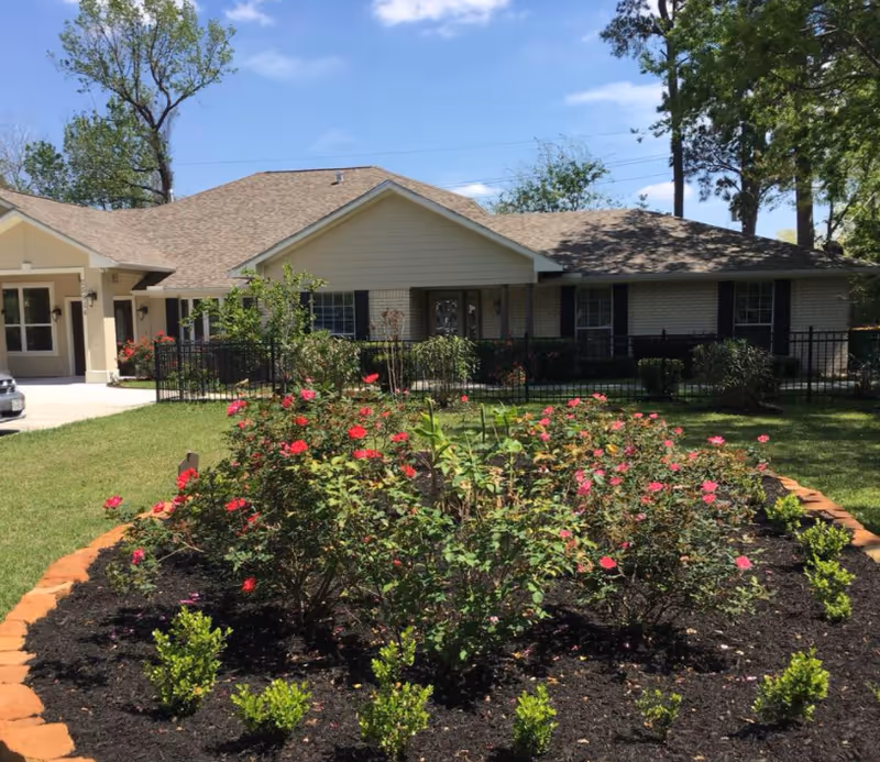 A single-story residential building with a beige exterior and a brown shingled roof, surrounded by a well-maintained garden with blooming red and pink flowers and green shrubs. The sky is clear and blue, and there are tall trees in the background.