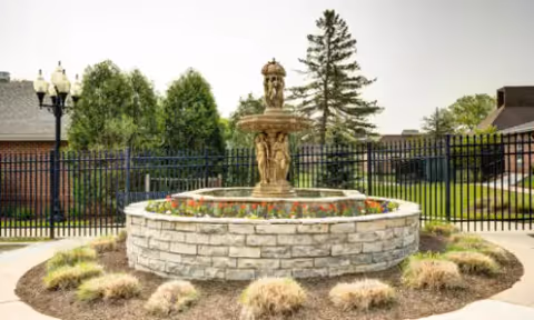 Stone tiered fountain in a circular stone planter with flowers and surrounding landscaping in a fenced courtyard.