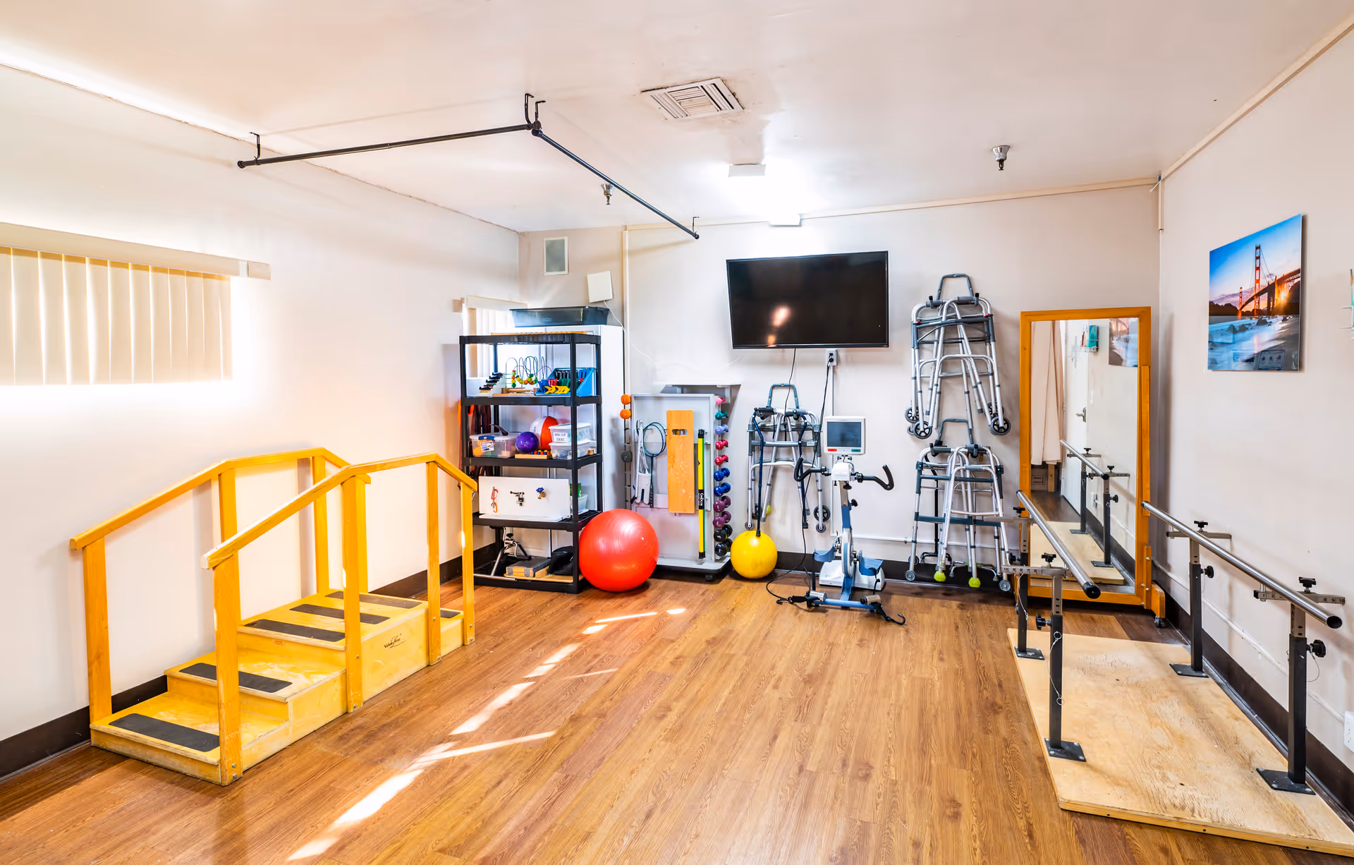 Bright therapy/exercise room with a small wooden practice staircase, parallel bars, exercise balls, walkers, and shelving with equipment.