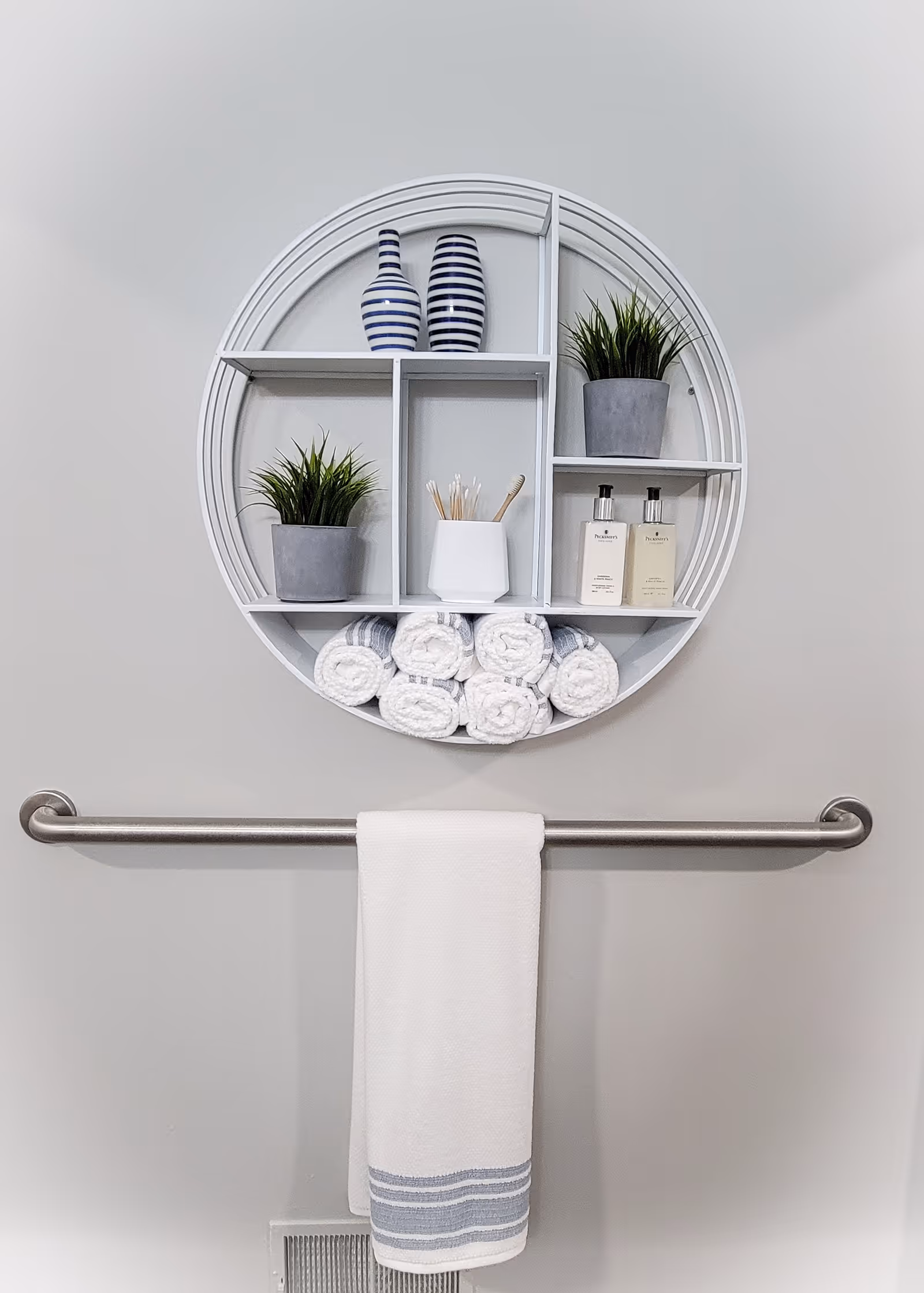 A round white wall shelf with multiple compartments holding two striped vases, two potted green plants, a white container with cotton swabs, two bottles of lotion or soap, and several rolled white towels. Below the shelf is a metal towel bar with a white towel hanging on it, featuring blue stripes at the bottom.
