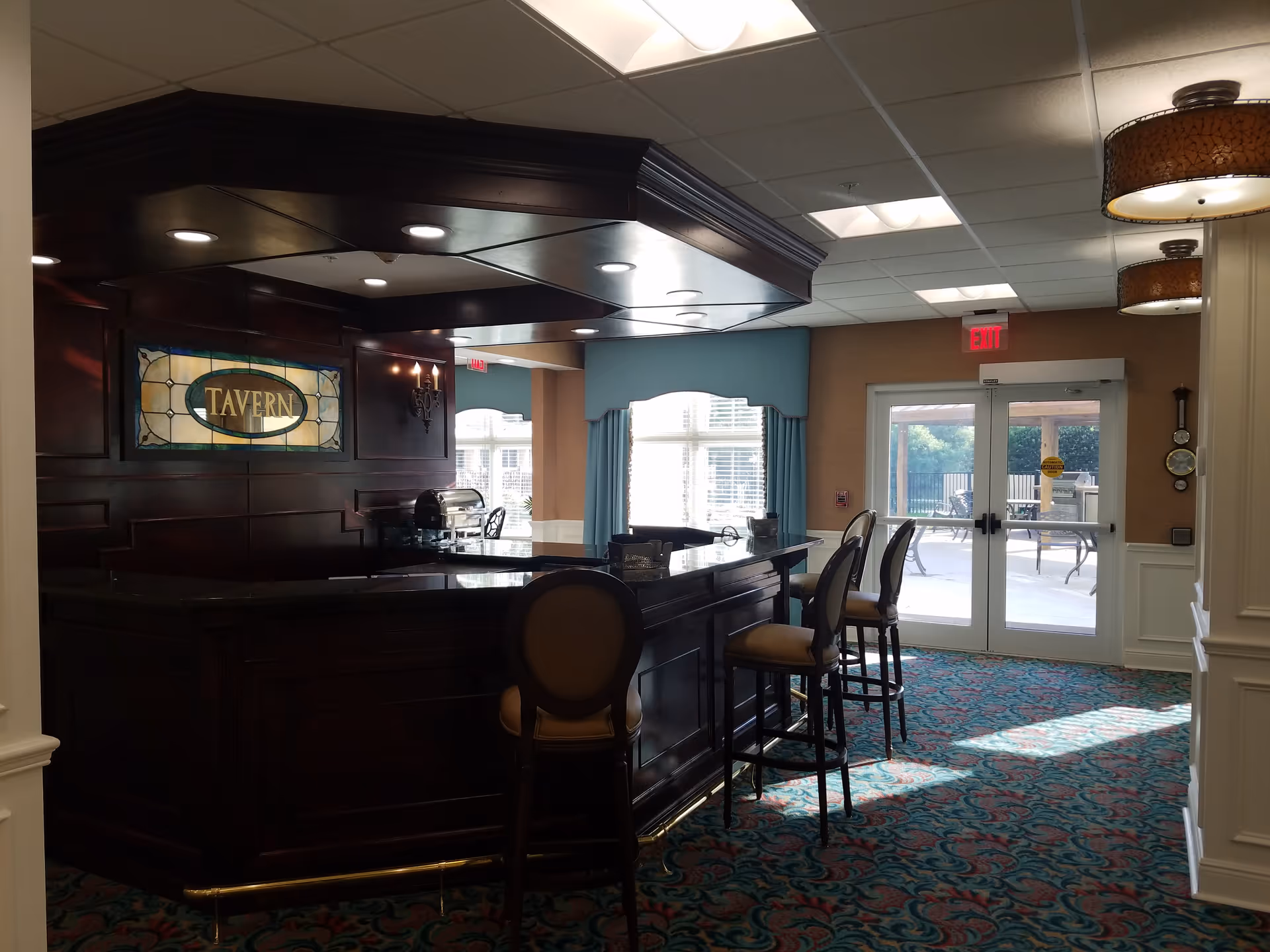 Interior view of a tavern area in a senior living facility with a dark wood bar counter, several high chairs, a stained glass sign that reads 'TAVERN', patterned carpet, and glass doors leading to an outdoor patio with tables and chairs.