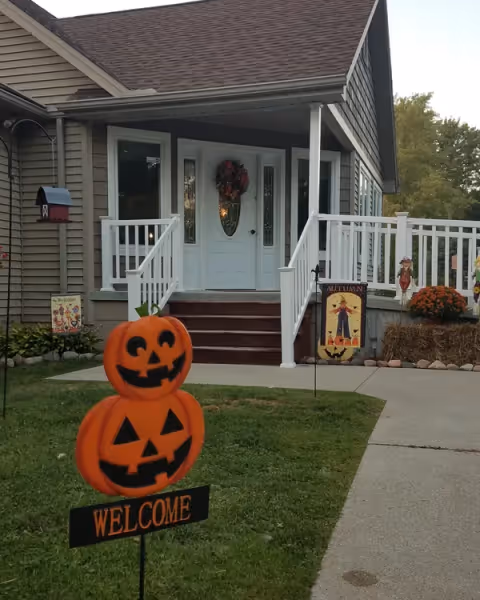 Front entrance of a house with a white porch and Halloween decorations, including a stacked jack-o'-lantern 'Welcome' sign on the lawn.