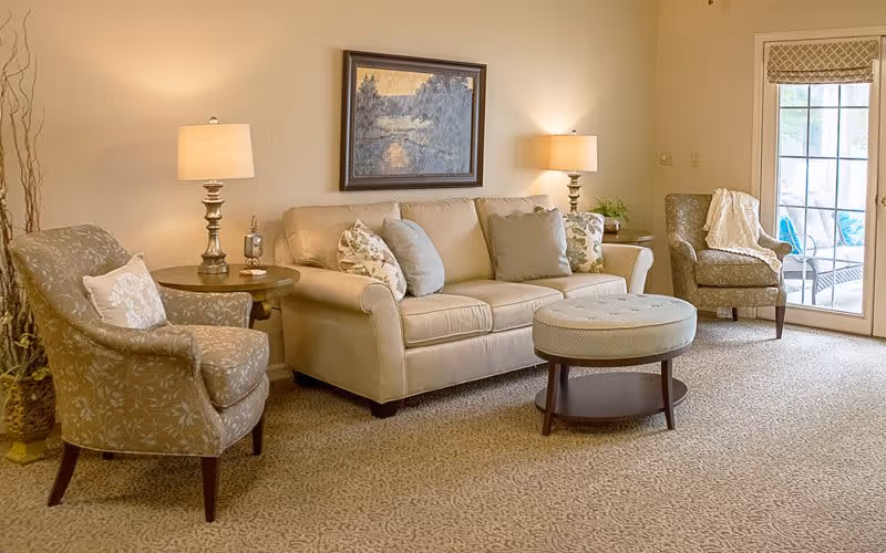 A cozy living room with a beige sofa adorned with multiple cushions, two patterned armchairs, a round ottoman coffee table, two table lamps on side tables, a framed landscape painting on the wall, and a glass door leading to an outdoor patio area.