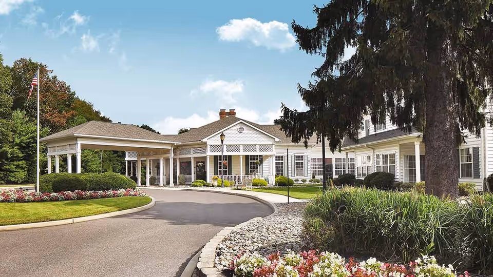 Front exterior view of a senior living facility named The Residence at Voorhees, featuring a circular driveway, a covered entrance, well-maintained landscaping with flowers, bushes, and trees, and an American flag on a flagpole under a partly cloudy sky.