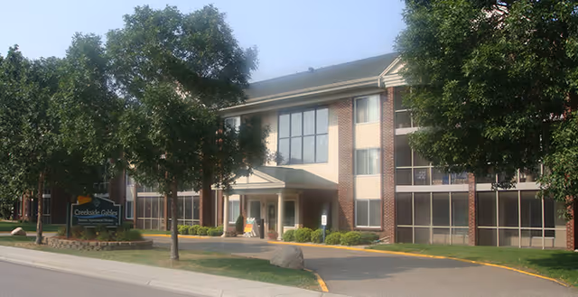 Exterior view of Creekside Gables, a two-story senior living facility with brick and beige siding, large windows, a covered entrance, and trees in front along a driveway.