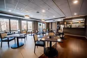 A spacious dining room with multiple square tables and black chairs arranged neatly. Large windows on one side allow natural light to fill the room, and there is a counter area at the back with shelves and decorative items. The floor is wooden, and the ceiling has recessed lighting.