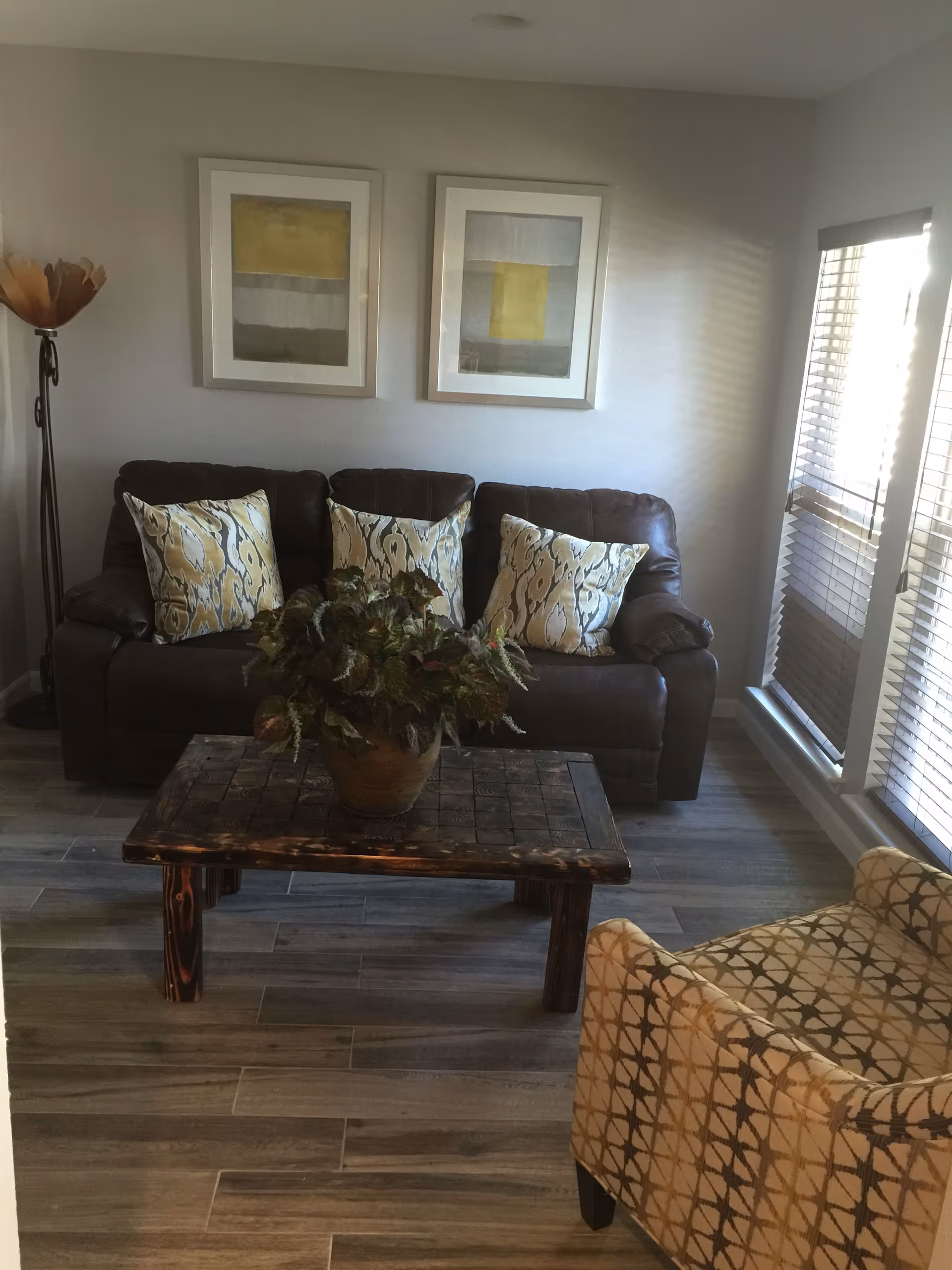 A cozy living room with a dark brown leather sofa adorned with three patterned cushions, a wooden coffee table with a potted plant on top, a patterned armchair in the foreground, two abstract paintings on the wall, a tall floor lamp in the corner, and large windows with blinds letting in natural light.