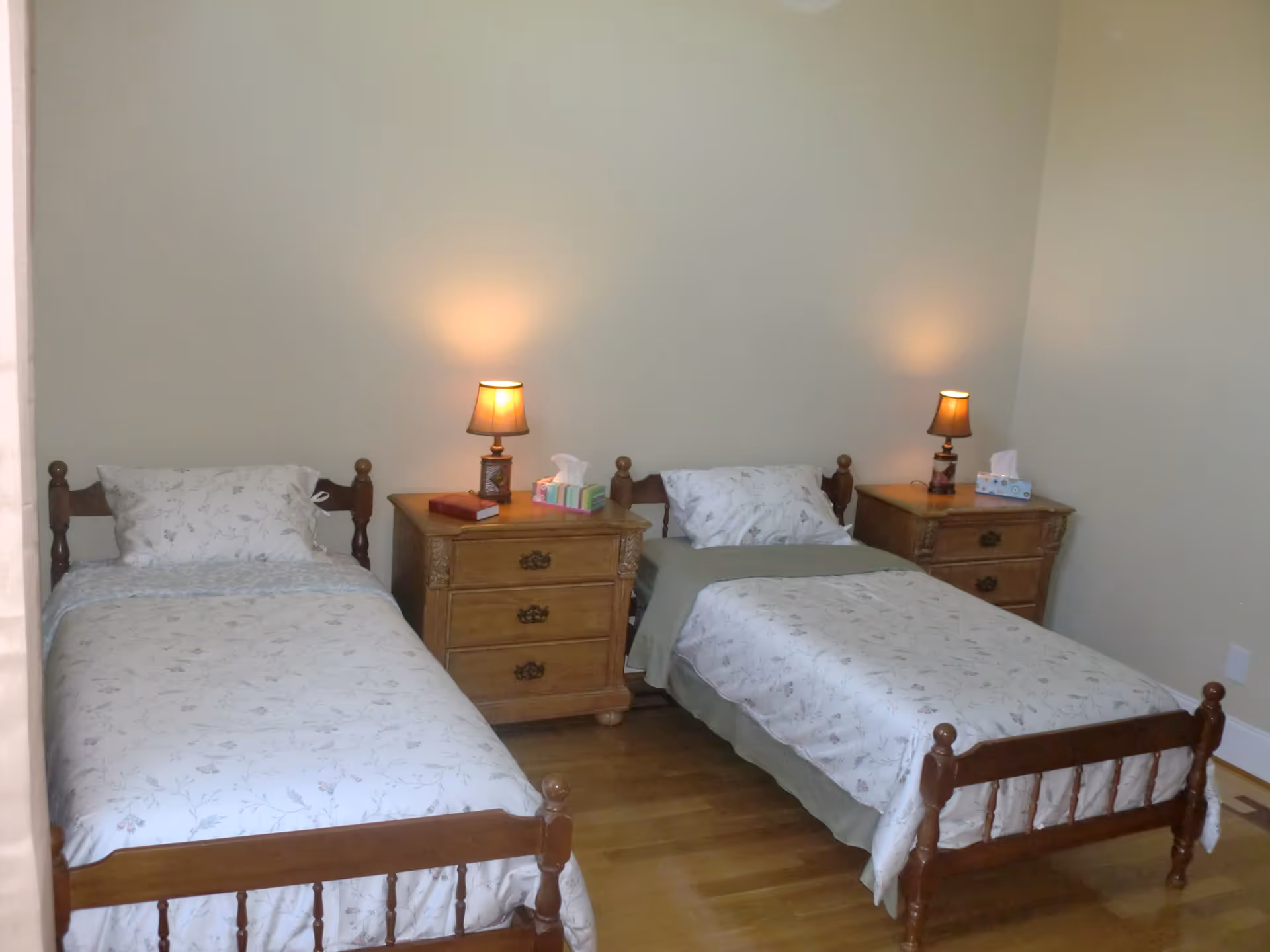 A simple bedroom with two single wooden beds placed parallel to each other, each with floral-patterned bedding and pillows. Between the beds are two wooden nightstands, each with a small lamp and a box of tissues. The room has plain light-colored walls and a wooden floor.