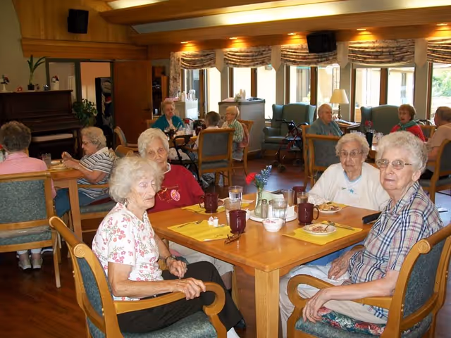 A group of elderly women sitting around a wooden dining table in a well-lit room with large windows. The table has yellow placemats, cups, plates with food, and a small flower vase. Other elderly individuals are seated at tables in the background, and the room has wooden floors and furniture, with a piano visible on the left side.