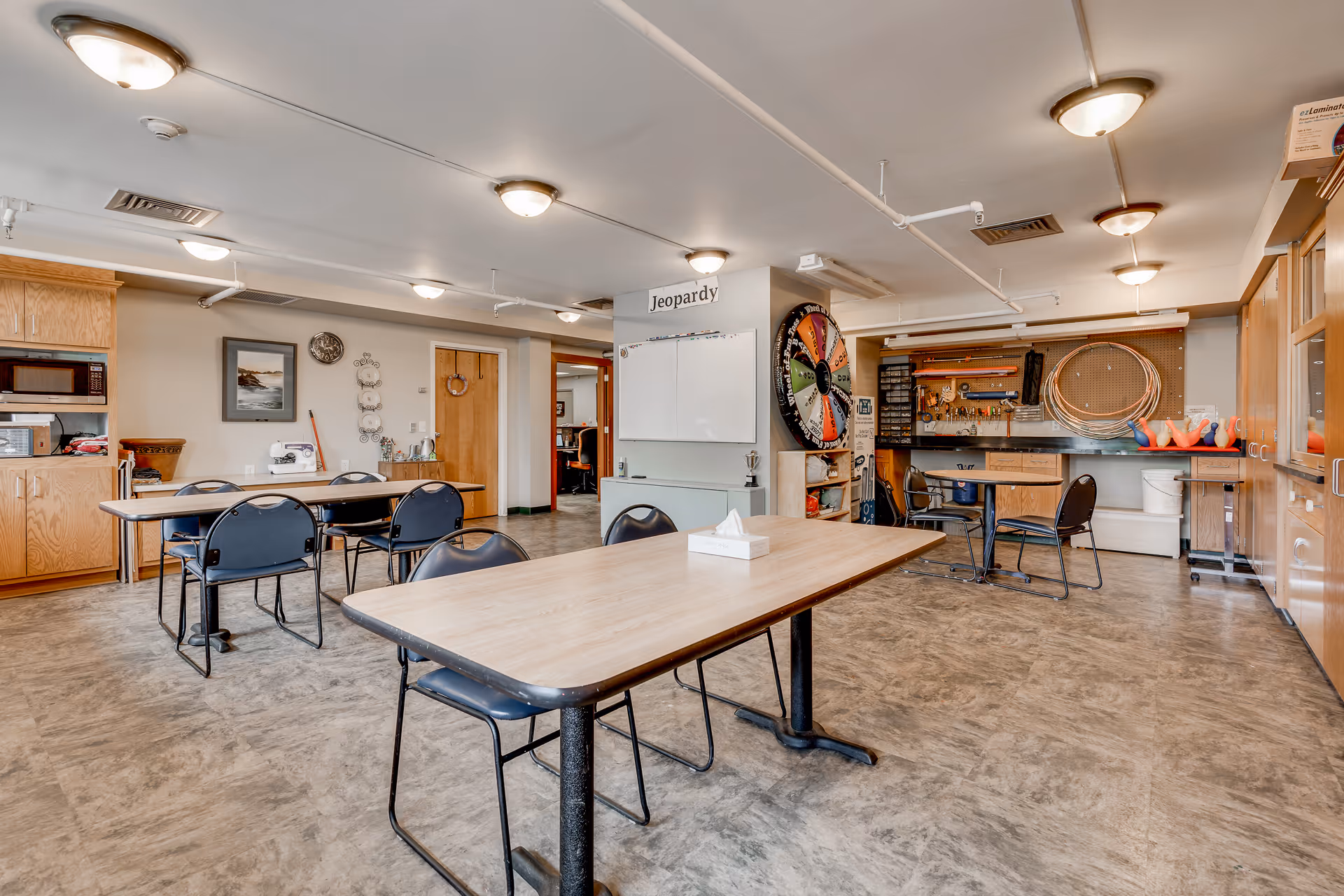 Activity room with long tables and chairs, a whiteboard labeled 'Jeopardy', and shelves of games and equipment along the walls.
