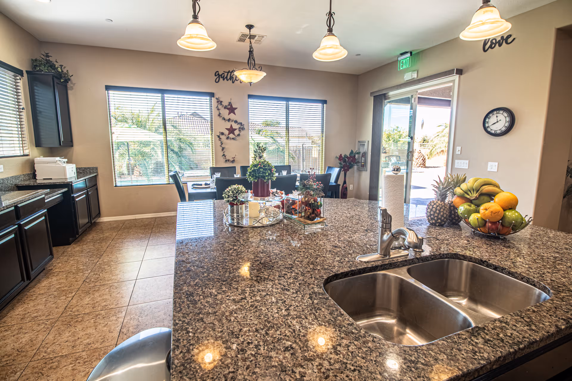 Bright kitchen and dining area with granite countertops, a double sink, and a bowl of assorted fruits on the counter. The dining table is set with chairs and decorative plants. Large windows with blinds let in natural light, and there are pendant lights hanging from the ceiling. The walls have decorative words 'gather' and 'love'. A sliding glass door leads to an outdoor area with palm trees visible.