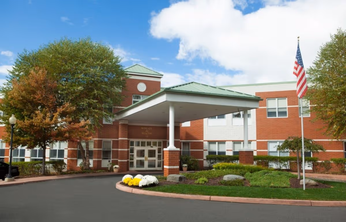 Front entrance of a brick senior living building with a covered porte-cochere, landscaping, and an American flag.