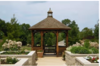 A wooden gazebo with a shingled roof situated in a garden area surrounded by green trees and flowering plants, with stone walls and planters on either side of the pathway leading to the gazebo.