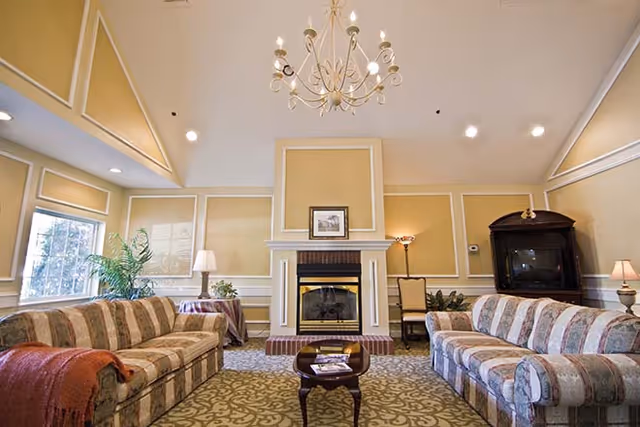 A spacious living room with two patterned sofas facing each other, a small wooden coffee table in the center, a fireplace with a framed picture above it, a chandelier hanging from the ceiling, and large windows letting in natural light. There are also several lamps and plants decorating the room.
