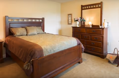 A well-lit bedroom featuring a wooden bed with pillows and a matching dresser topped with a mirror and decorative items.