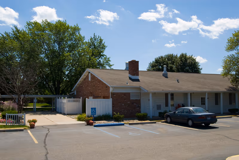 Single-story brick senior living building with a parking lot, a parked car, and trees under a blue sky.