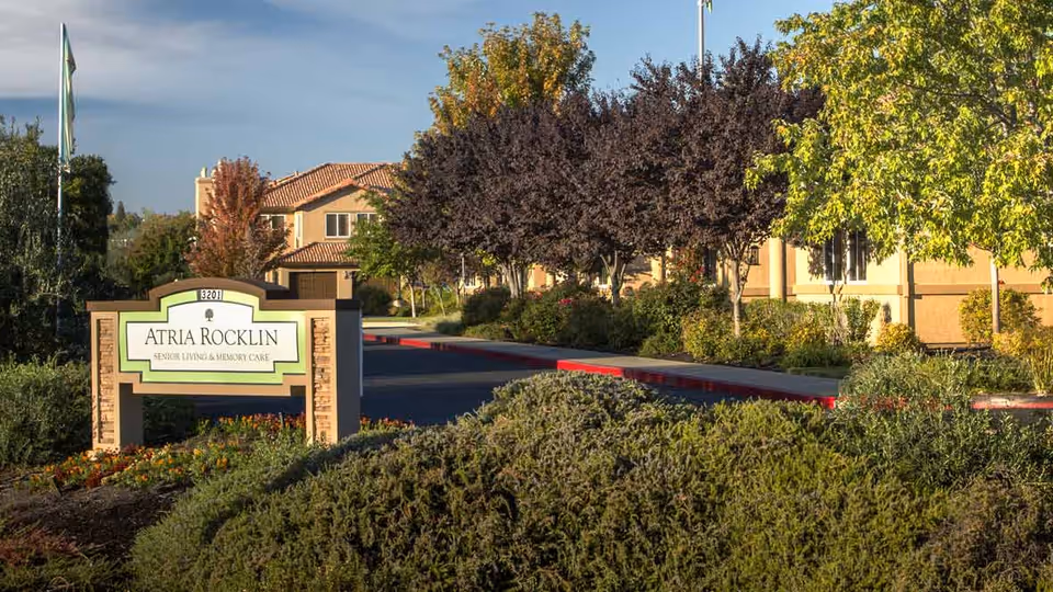 Entrance sign for Atria Rocklin senior living and memory care facility surrounded by landscaped bushes and trees with a building in the background under a clear sky.