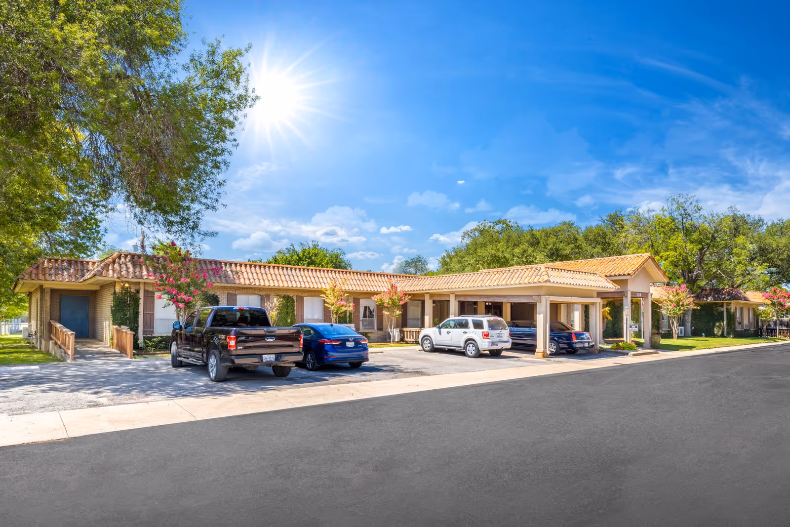 Exterior view of 214 Martin-A Retirement Residence on a sunny day with a clear blue sky. The building has a tiled roof and beige brick walls, with several cars parked in front. There are trees and flowering plants around the building.