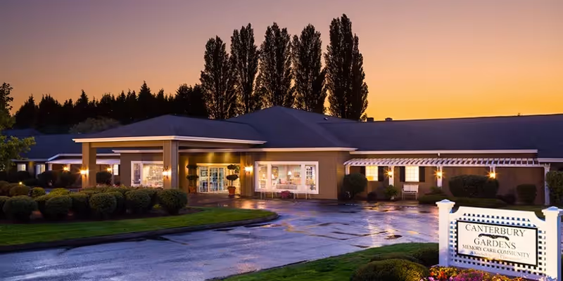 Exterior view of Canterbury Gardens Memory Care Community building at sunset with illuminated entrance, well-maintained landscaping, and tall trees in the background.