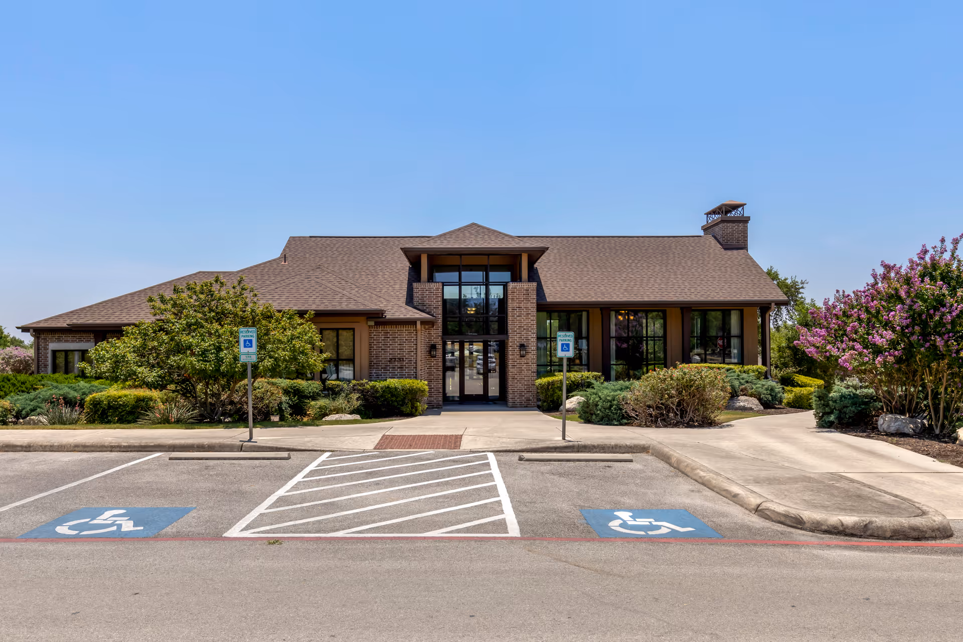 Front exterior of a single-story brick senior living facility with a central glass entrance, landscaping, and marked handicap parking spaces.