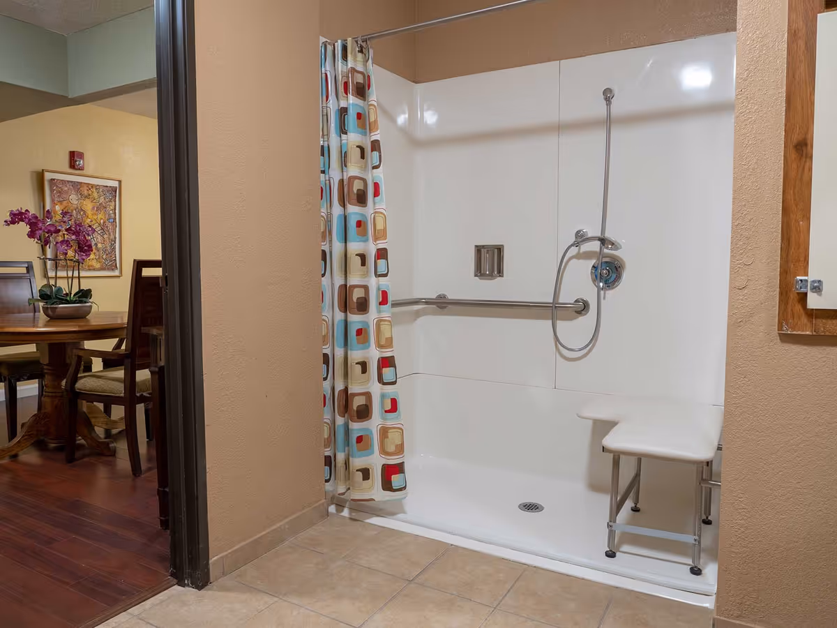 A walk-in shower with a white foldable shower seat and a colorful shower curtain with a geometric pattern. The shower has a handheld showerhead and a grab bar for safety. Adjacent to the shower is a view into a dining area with a wooden table, chairs, a purple orchid centerpiece, and a framed artwork on the wall.