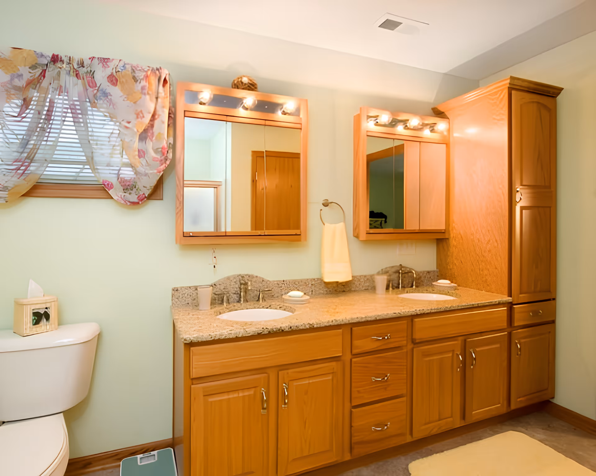 A bathroom with a double sink vanity featuring wooden cabinets and granite countertop. Above each sink is a wooden framed mirror with lights. To the left is a toilet with a tissue box on top and a window with floral curtains. A yellow hand towel hangs between the two mirrors.