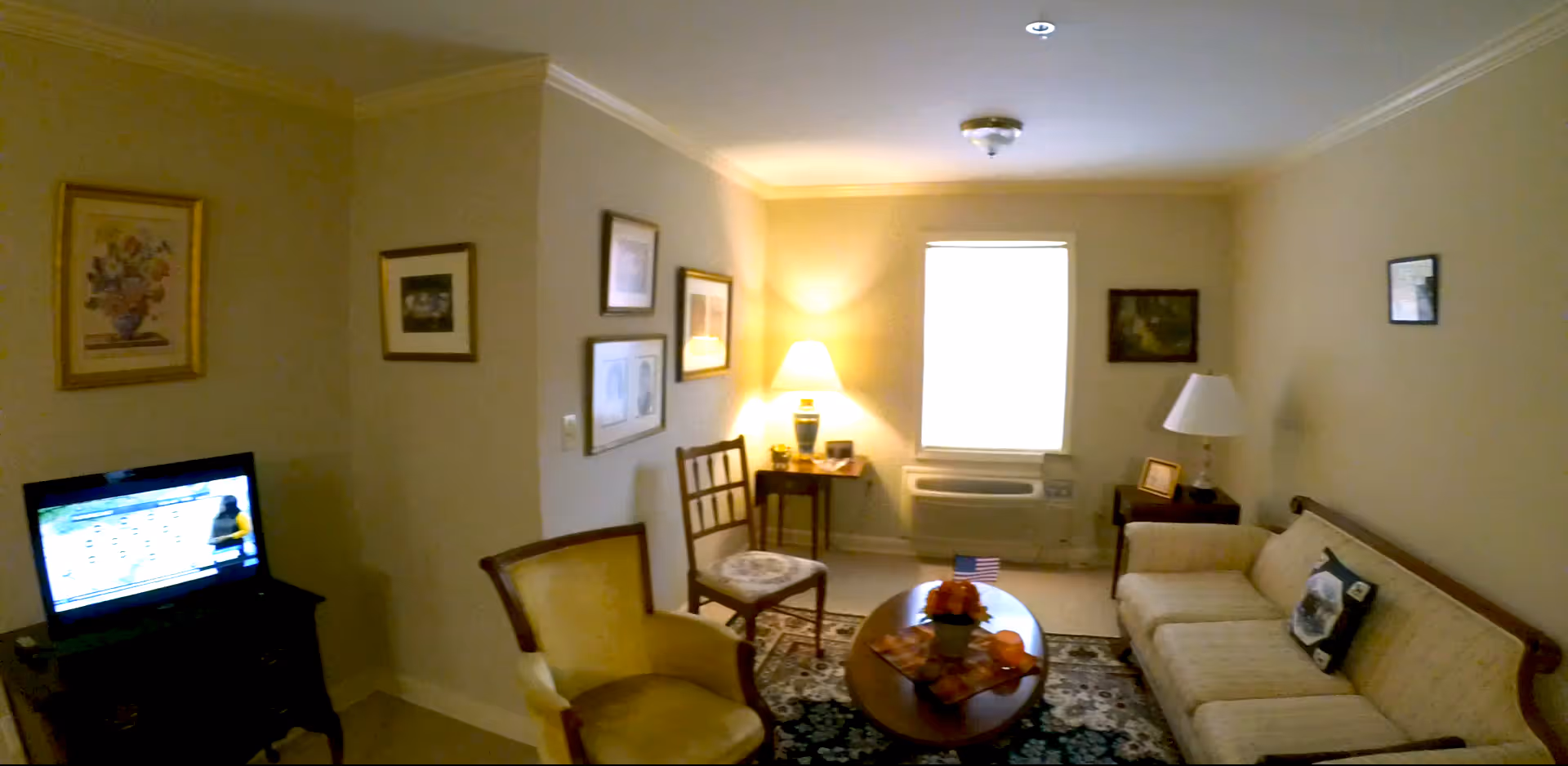 A cozy living room with a beige sofa, a wooden coffee table with a small centerpiece, two chairs, a TV on a stand, framed pictures on the walls, a window with a white blind, and two table lamps providing warm lighting.