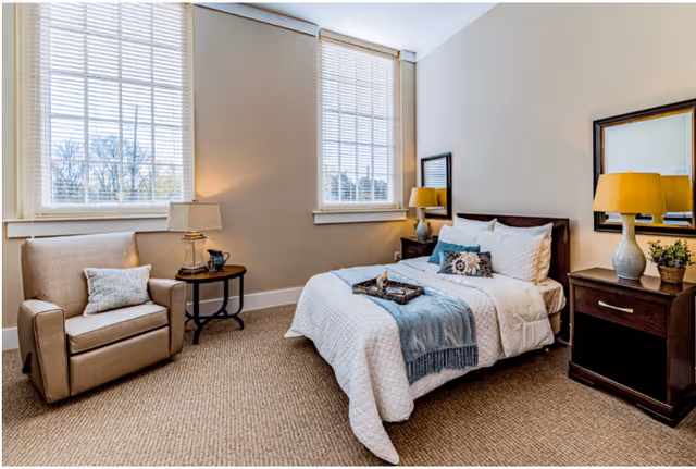 A cozy bedroom with a neatly made bed featuring white bedding and a blue throw blanket. There is a beige armchair with a pillow next to a small round side table with a lamp and decorative items. Two large windows with blinds allow natural light to fill the room. On the right side, there is a dark wooden nightstand with a yellow lamp and a small plant, and a mirror hanging above it.