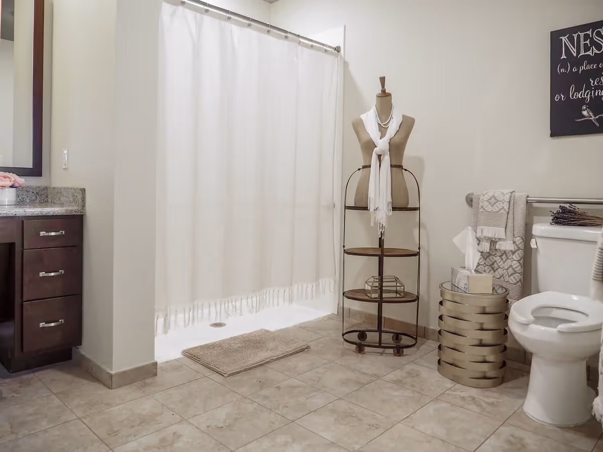 A bathroom with a white shower curtain, beige tiled floor, a toilet with a towel rack above it holding folded towels, a decorative mannequin stand with a white scarf, a small round side table with a tissue box, and a dark wood vanity with granite countertop and drawers.