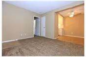 Empty room with beige carpet and beige walls, a white door slightly open leading to another room, and an adjacent area with tiled flooring and a ceiling fan with light.