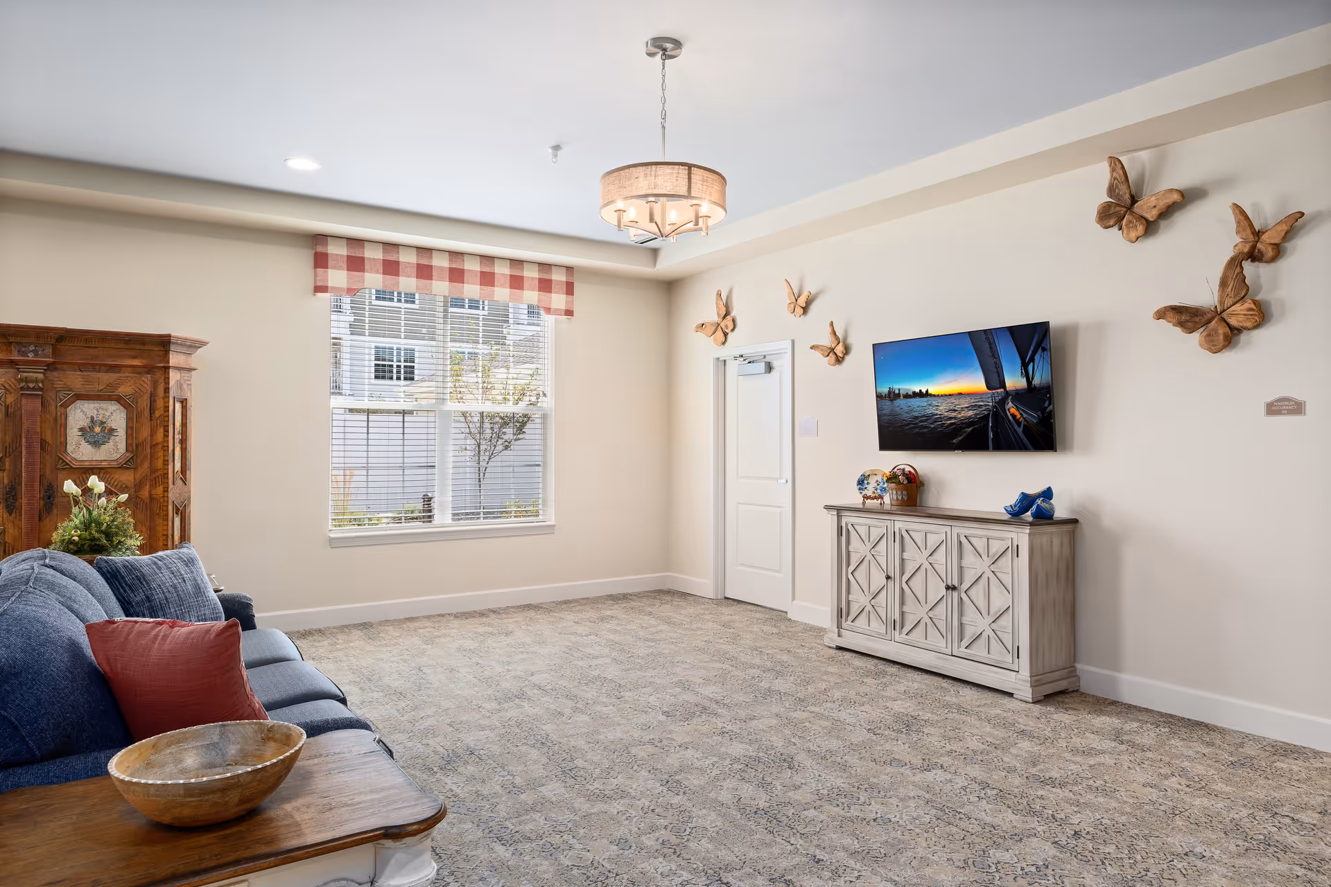 A bright and spacious living room with a blue sofa adorned with red and blue pillows, a wooden side table with a decorative bowl, a large window with a red and white checkered valance, a wooden cabinet with floral decorations, a wall-mounted flat-screen TV displaying a sunset over water, and wooden butterfly wall decorations.