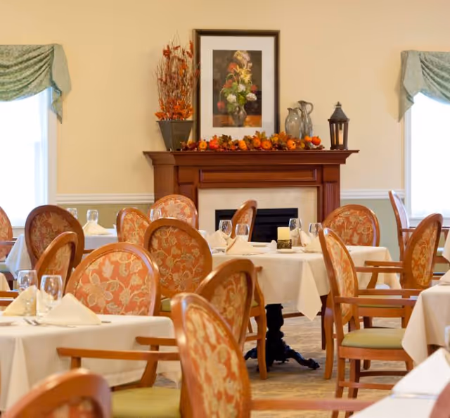Dining room with tables covered in white tablecloths, set with napkins and glassware. The room features wooden chairs with floral upholstery, two windows with green valances, and a fireplace mantel decorated with a floral arrangement, a framed flower painting, and autumn-themed decor.