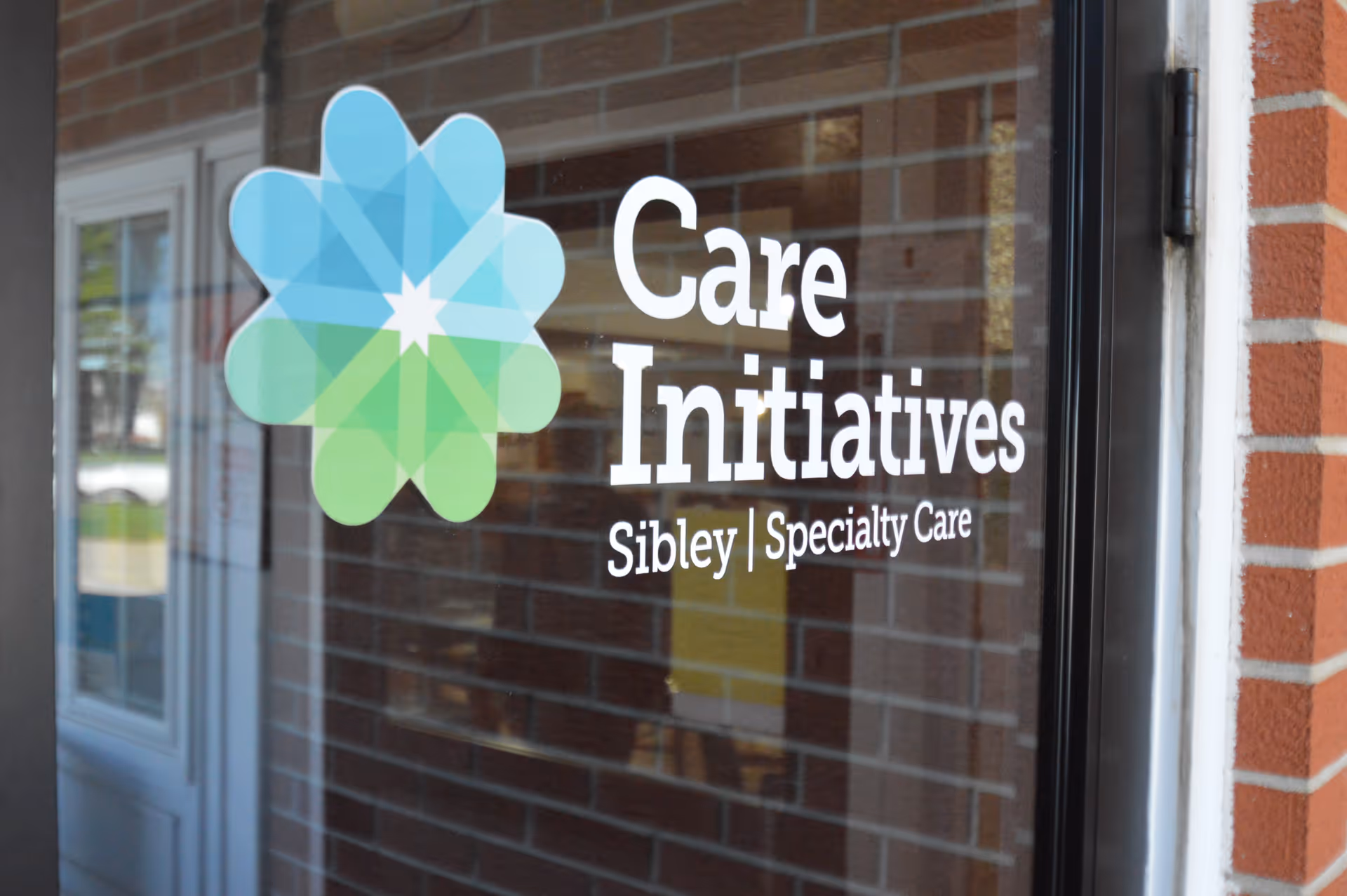 Glass entrance door with a green-blue flower logo and white text reading 'Care Initiatives Sibley | Specialty Care' against a brick background.