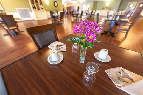A dining area in a senior living facility with wooden tables and chairs. The foreground table has a vase with purple flowers, two cups and saucers, a glass turned upside down, and a napkin with cutlery. The background shows more tables and chairs with soft lighting and a warm, inviting atmosphere.