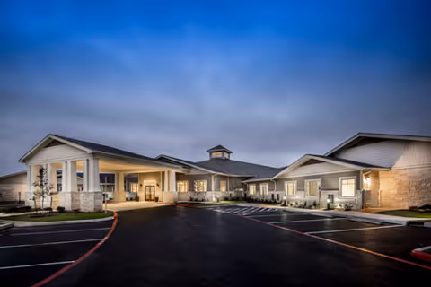 Front exterior of a single-story senior living facility at dusk with a covered lit entrance and empty parking area.