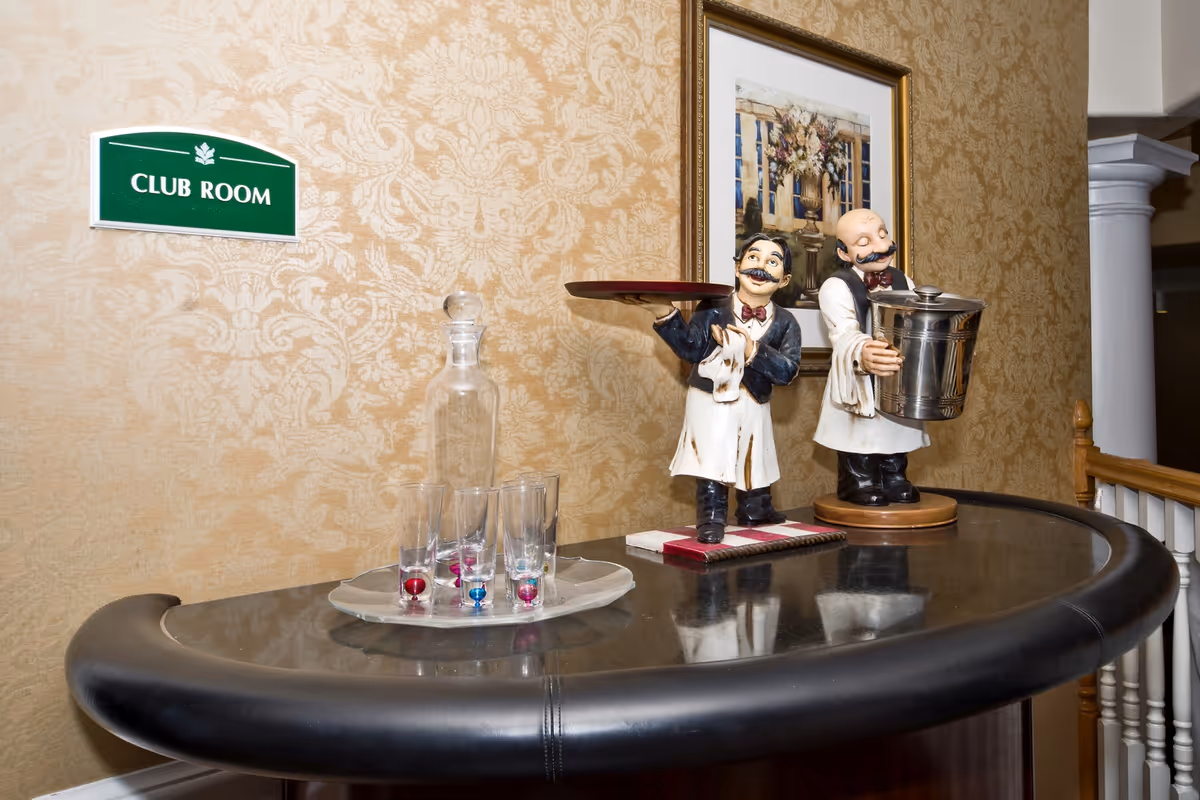 A curved black bar counter with two ceramic waiter figurines, one holding a tray and the other holding an ice bucket. On the counter, there is a glass decanter and four glasses with colored marbles inside on a tray. A framed floral painting hangs on the patterned wallpapered wall behind the counter. A green sign on the wall reads 'CLUB ROOM'.