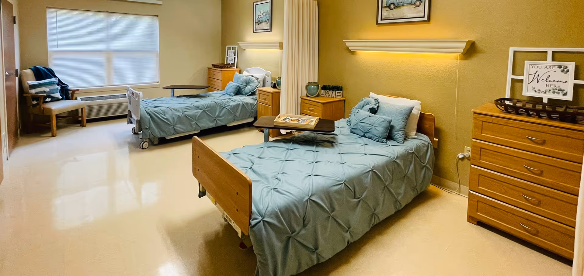 A well-lit room with two single hospital-style beds, each with teal bedding and pillows. There are wooden nightstands and dressers beside the beds, a chair near a large window with blinds, and a small table on wheels positioned over one of the beds. The walls are painted a warm beige color and decorated with framed pictures and a sign that reads 'You are Welcome Here.'