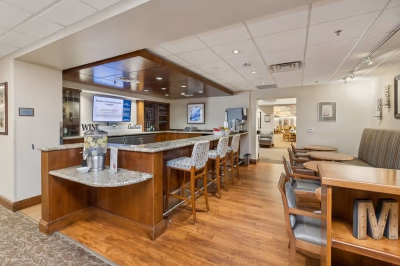 Interior view of a senior living facility's dining and bar area featuring a wooden bar counter with granite top, four cushioned bar stools, a water dispenser with lemon water, and several wooden tables with chairs along the right wall. The space has wood flooring, framed artwork on the walls, and a ceiling with recessed lighting. A TV screen is mounted behind the bar.