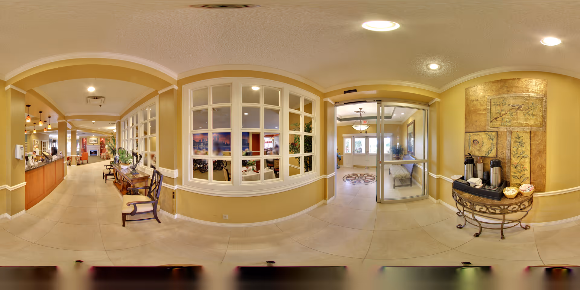 Interior view of a senior living facility lobby area with a reception desk on the left, seating area visible through large windows in the center, and a small table with coffee and condiments on the right. The walls are painted yellow with white trim, and the floor is tiled. There are decorative light fixtures and artwork on the walls.