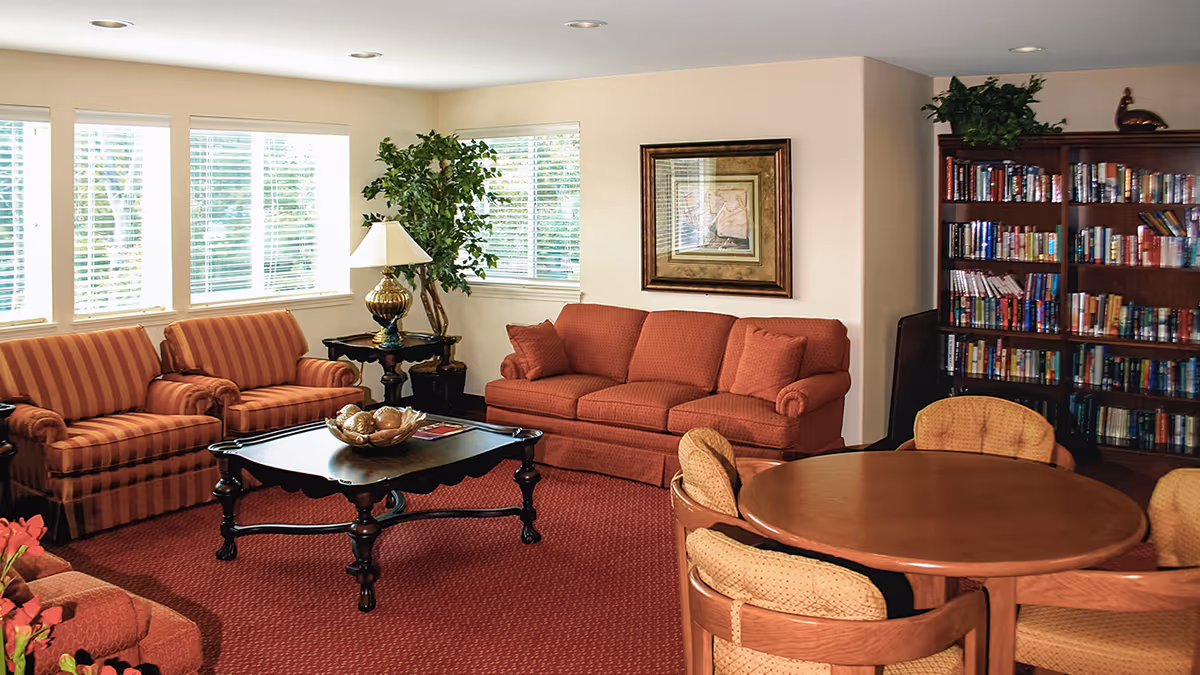 A cozy living room with red carpet and matching red sofas, a wooden coffee table with decorative items, a round wooden table with cushioned chairs, a large bookshelf filled with books, a framed picture on the wall, a potted plant, and windows with white blinds letting in natural light.